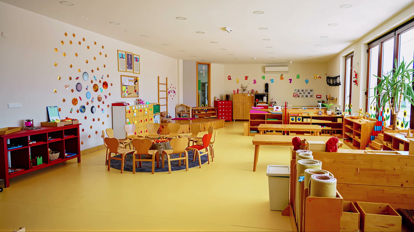 Geniş sınıf: çember masası, Montessori materyal rafları ve sarı zemin